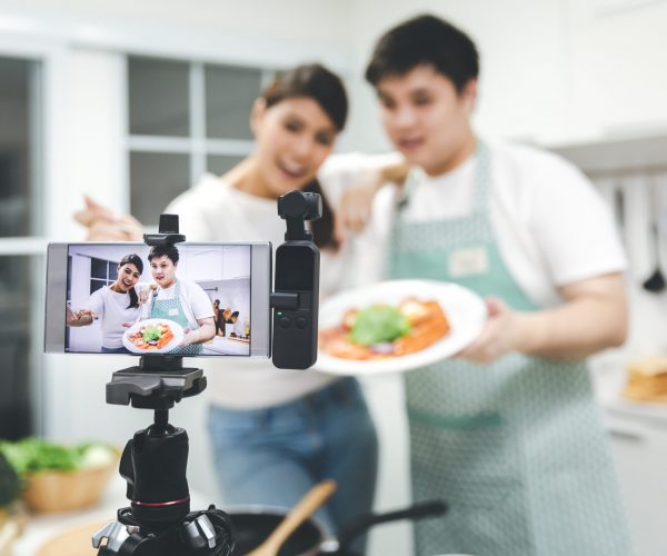 Vlogger and blogger freelance job concept, Young asian couple cooking together and recording live video for her vlog and social media with mobile phone camera and tripod